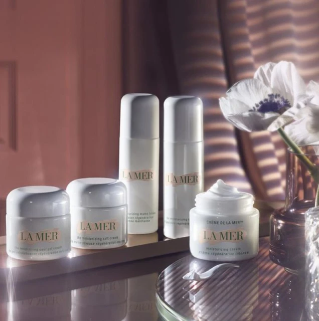 La Mer skincare products, including creams and lotions, are displayed on a reflective surface with a white flower in a vase.