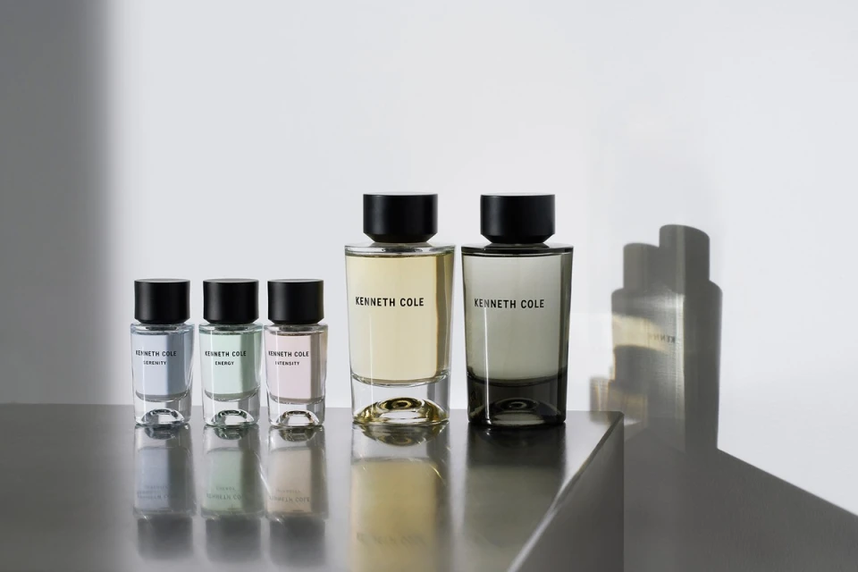 A row of Kenneth Cole fragrance bottles in varying sizes and colors on a reflective gray surface, set against a plain white background.