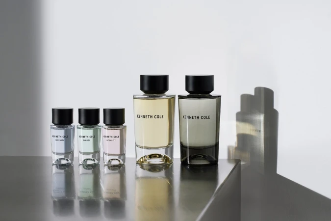 A row of Kenneth Cole fragrance bottles in varying sizes and colors on a reflective gray surface, set against a plain white background.