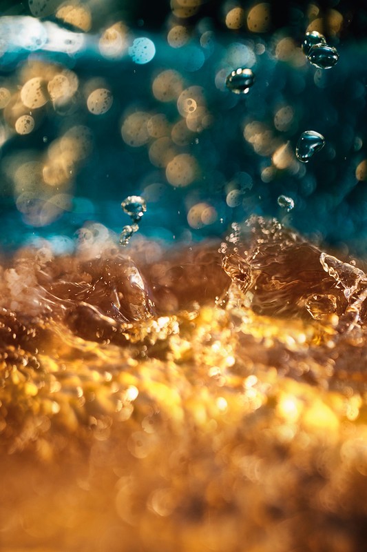 Close-up of water with an amber hue, featuring water droplets suspended in the air against a blurred teal and yellow background.