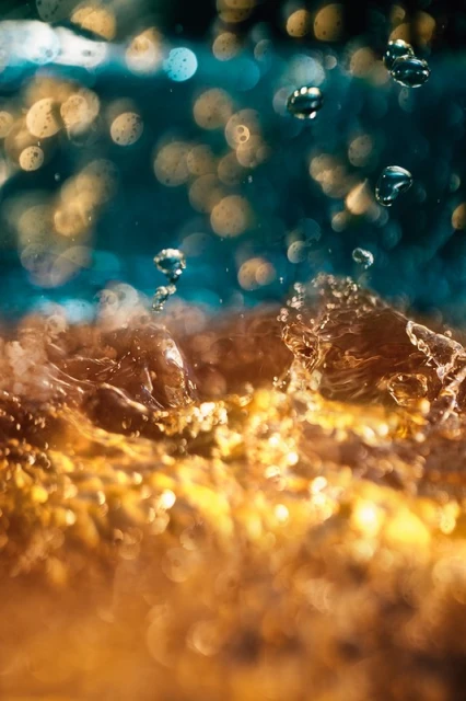 Close-up of water with an amber hue, featuring water droplets suspended in the air against a blurred teal and yellow background.