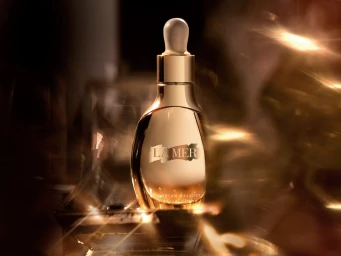 Close up product photography of a gold serum bottle with a dropper cap surrounded by warm glowing light flares.