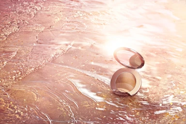 Round compact makeup powder case resting on wet sand near shallow water glistening in warm sunlight