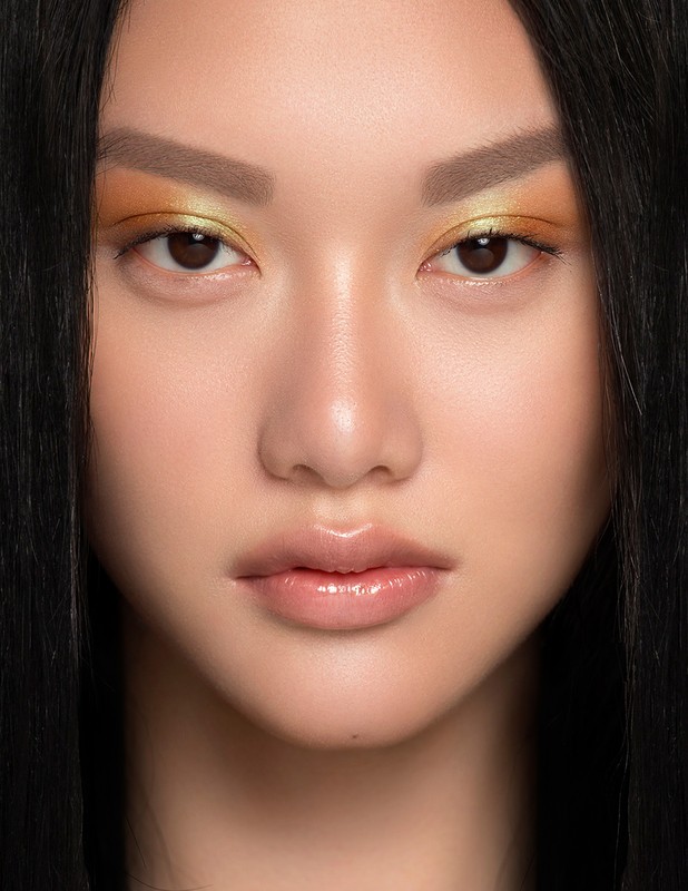Close-up portrait of a woman with vibrant yellow and orange eyeshadow, glossy lips, and dark hair framing her face.