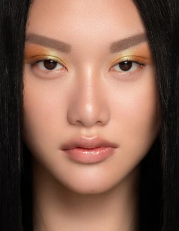 Close-up portrait of a woman with vibrant yellow and orange eyeshadow, glossy lips, and dark hair framing her face.