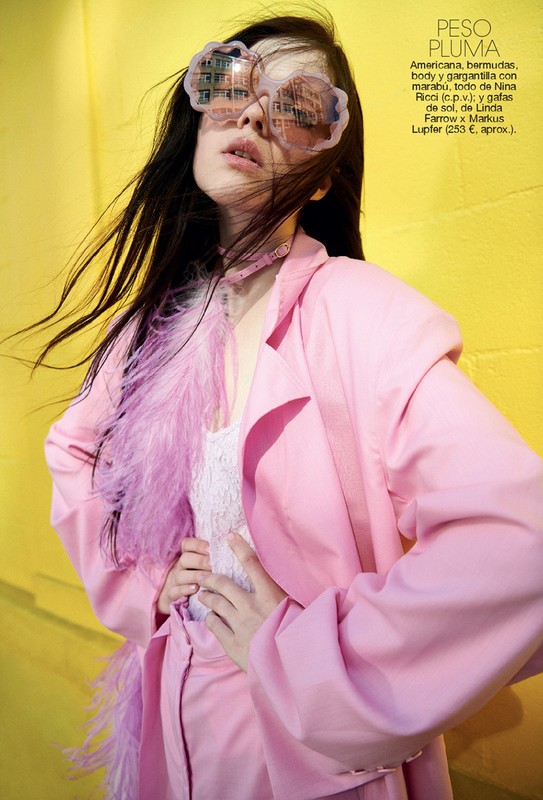Fashion shot of a woman in a pink suit and sunglasses, posed against a yellow wall, featuring feathered details and a stylish editorial look.