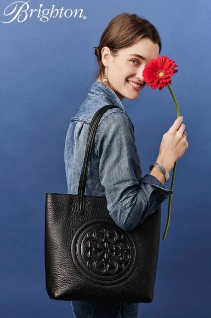 A woman wearing a denim jacket and holding a red flower stands next to a black leather tote bag in a fashion product shot.