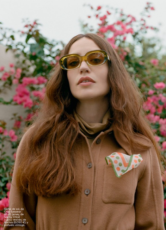 A young woman poses outdoors wearing sunglasses, a button-down jacket, and a scarf, with pink flowers and foliage in the background.