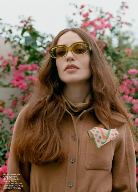 A young woman poses outdoors wearing sunglasses, a button-down jacket, and a scarf, with pink flowers and foliage in the background.