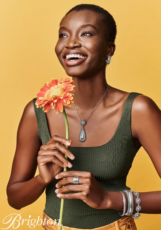Smiling woman poses in a fashion shot, wearing jewelry and holding an orange gerbera daisy against a bright yellow backdrop.