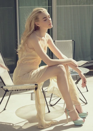 Blonde woman in a slip dress sits in the sun on a patio with metal chairs, wearing high heels and her eyes closed in relaxation.
