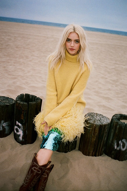 Blonde woman in yellow sweater and cowboy boots sits on driftwood on a sandy beach with the ocean in the background.