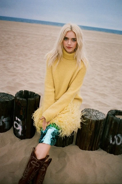 Blonde woman in yellow sweater and cowboy boots sits on driftwood on a sandy beach with the ocean in the background.
