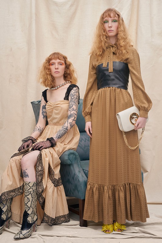 Two red-haired women model earth-toned vintage dresses against a neutral backdrop, one seated on a blue chair, the other standing.