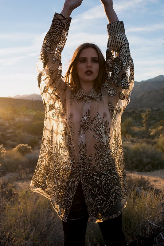Young woman with arms raised poses in a desert landscape, wearing a sheer embellished shirt against a backdrop of mountains and sky.