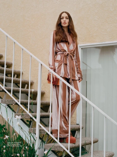 Woman in a striped pink pajama suit stands on stairs outdoors, holding an orange clutch, posing in front of a peach building.