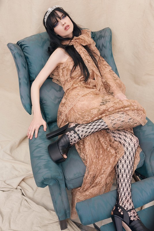 Woman in a tan lace gown reclines on a blue chair, wearing black platform heels and fishnet stockings against a neutral backdrop.