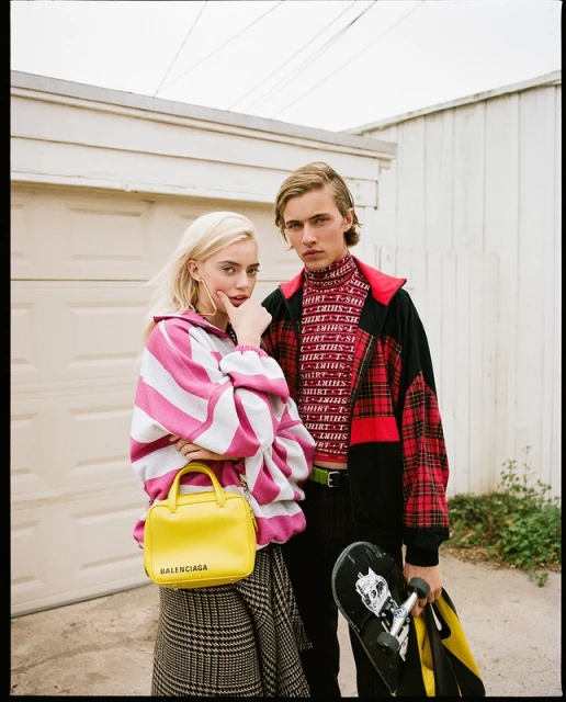 Two fashion models pose outdoors near a garage; the female model carries a yellow Balenciaga purse while the male model holds a skateboard.