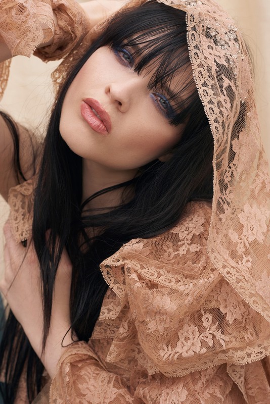 Close-up portrait of a fashionable young adult woman with black hair and bangs, wearing lace fabric over her head and shoulders.