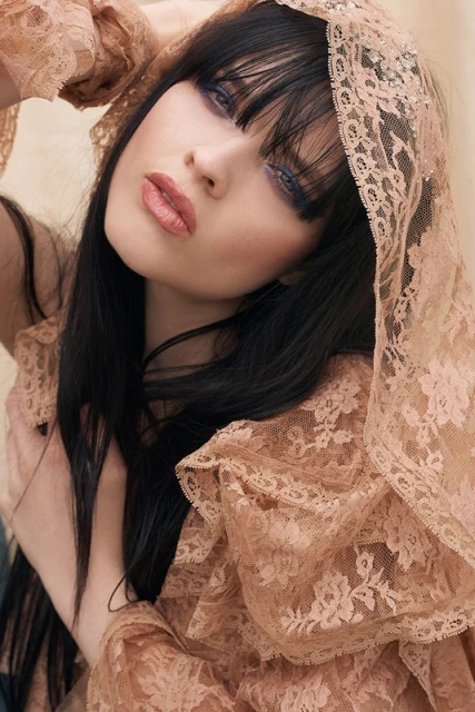 Close-up portrait of a fashionable young adult woman with black hair and bangs, wearing lace fabric over her head and shoulders.