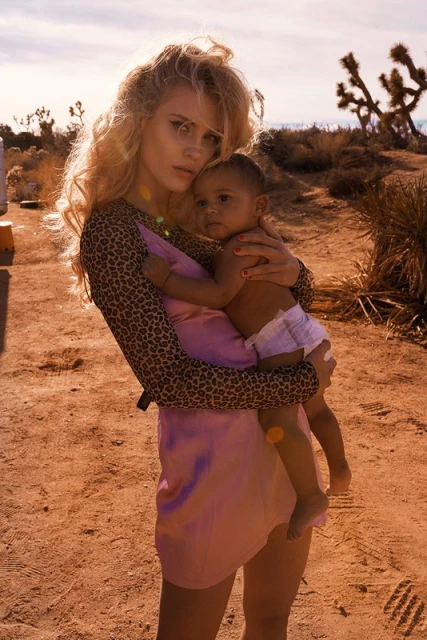 Woman holds baby in desert landscape, wearing a pink dress and animal print top with Joshua Trees in the background on a sunny day.