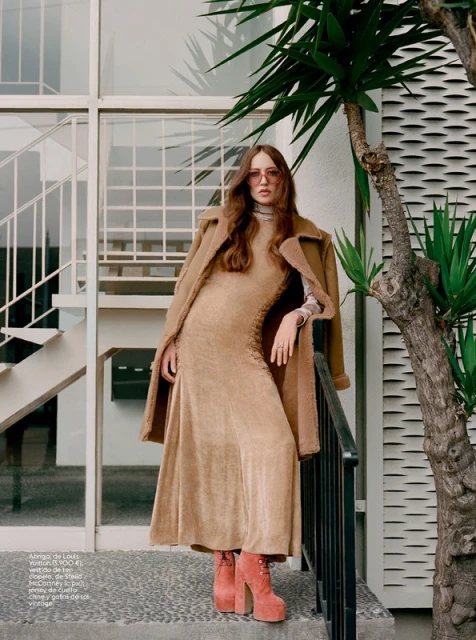 Woman in a neutral dress and coat posing outdoors on concrete steps with platform boots, next to a palm tree and glass-lined building.