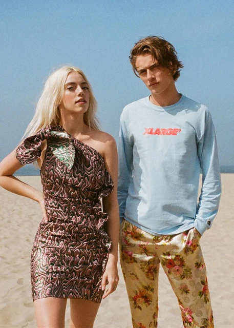 A couple poses on a sandy beach under a bright sky. The woman wears a ruffled dress, while the man is in a logo shirt and floral pants.
