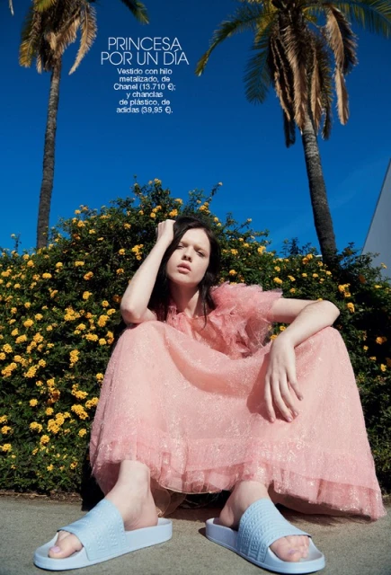 Brunette woman poses in a pink dress with palm trees against a blue sky. She's wearing light blue sandals for a fashion editorial.