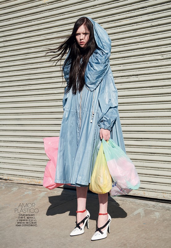 A young woman stands in a light blue hooded coat holding shopping bags, set against a gray shutter door and sunlit concrete.