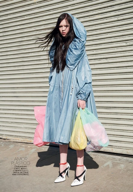 A young woman stands in a light blue hooded coat holding shopping bags, set against a gray shutter door and sunlit concrete.