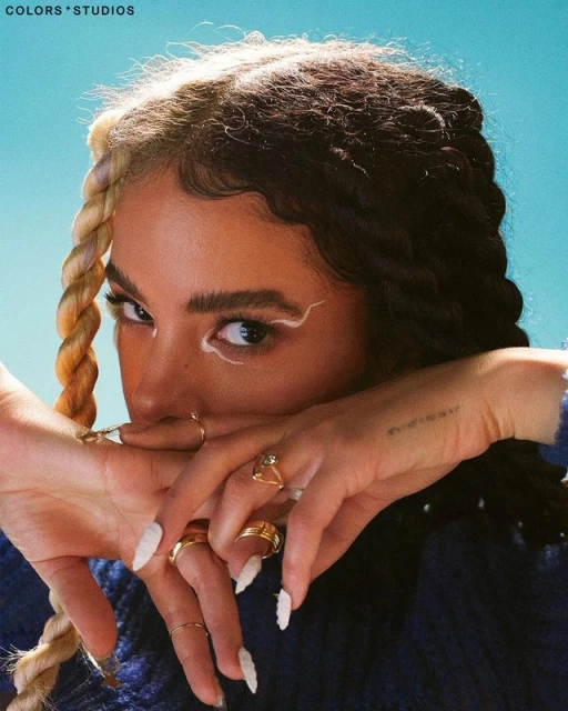 Close-up portrait of a woman with unique braided hair and white eyeliner posing in front of a solid light blue background.