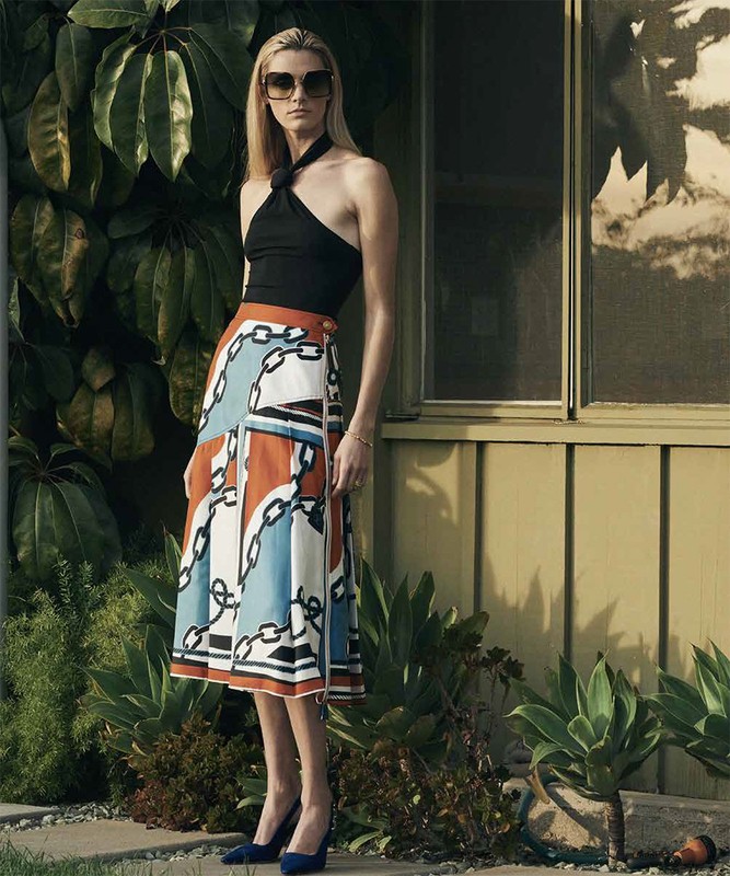 Full-length shot of a woman posing in a black halter top and patterned skirt with architectural and foliage background, outdoors.