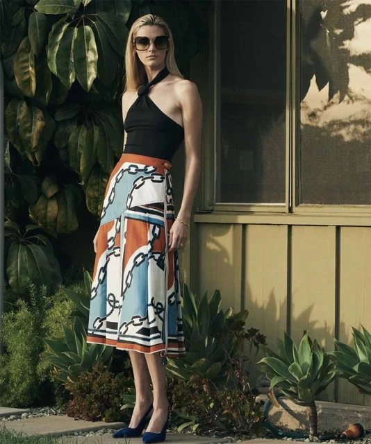 Full-length shot of a woman posing in a black halter top and patterned skirt with architectural and foliage background, outdoors.