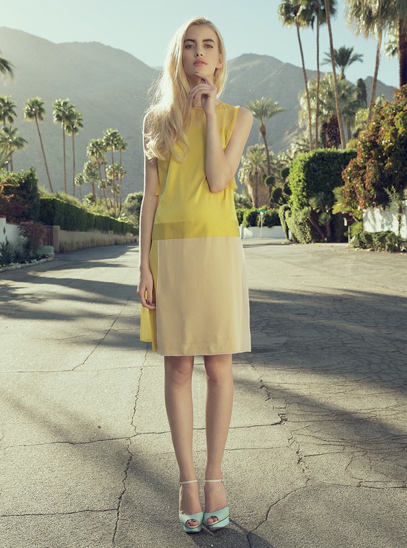 Fashion shot of a blonde model wearing a sleeveless yellow and beige dress and high heels, posing on a cracked asphalt road with mountains.