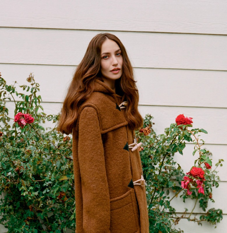 A young woman with long brown hair wears a brown wool coat and stands outside near a white wall and flowering rose bushes.