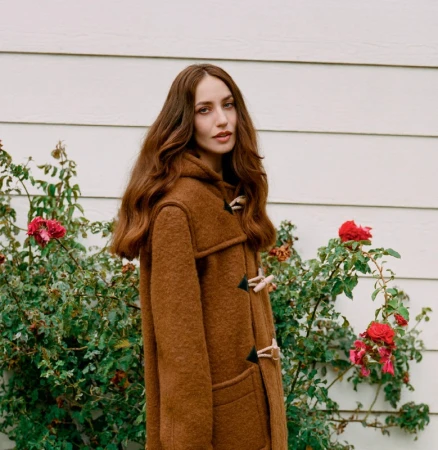 A young woman with long brown hair wears a brown wool coat and stands outside near a white wall and flowering rose bushes.