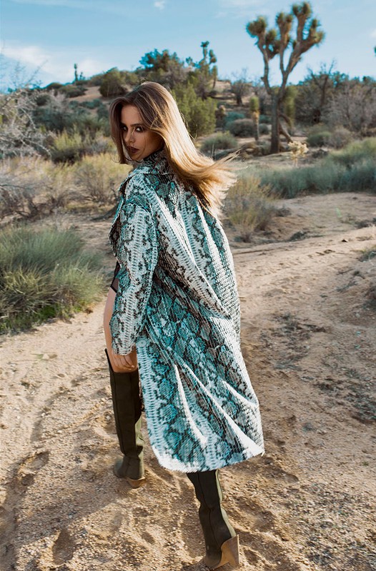 A woman in a snakeskin-patterned coat and thigh-high boots walks away on a desert trail, glancing over her shoulder at the camera.