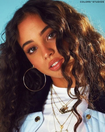Portrait of a young woman with long curly brown hair wearing hoop earrings and layered necklaces against a blue background.