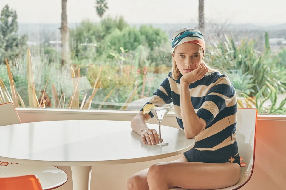 Blonde woman in a striped top sits at a white table, looking at the camera with a serious expression; a cocktail with a lemon wedge in it sits on the table.