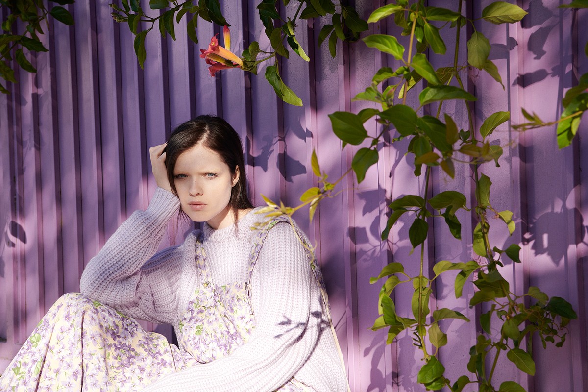 Young adult woman in a lavender sweater and floral dress poses outdoors against a purple fence with climbing green foliage and sunlight.