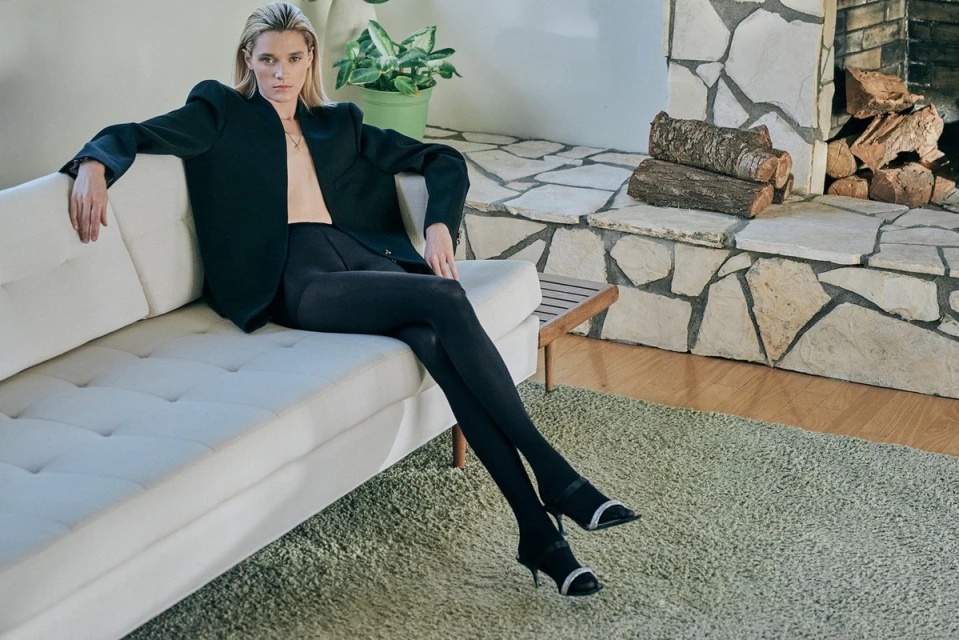 Woman in black blazer and leggings poses on a white couch in a modern interior with fireplace, plant, and carpet.