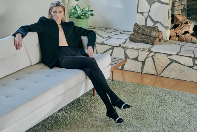 A blonde female model wearing a dark blazer and tights seated on a white sofa indoors with a stone fireplace.