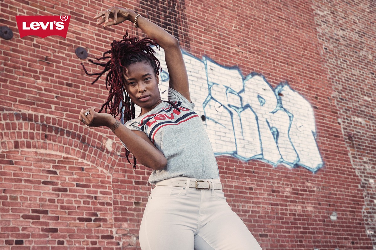 Young woman poses against a brick wall in a Levi's T-shirt and white jeans, showcasing fashion in an urban outdoor setting.