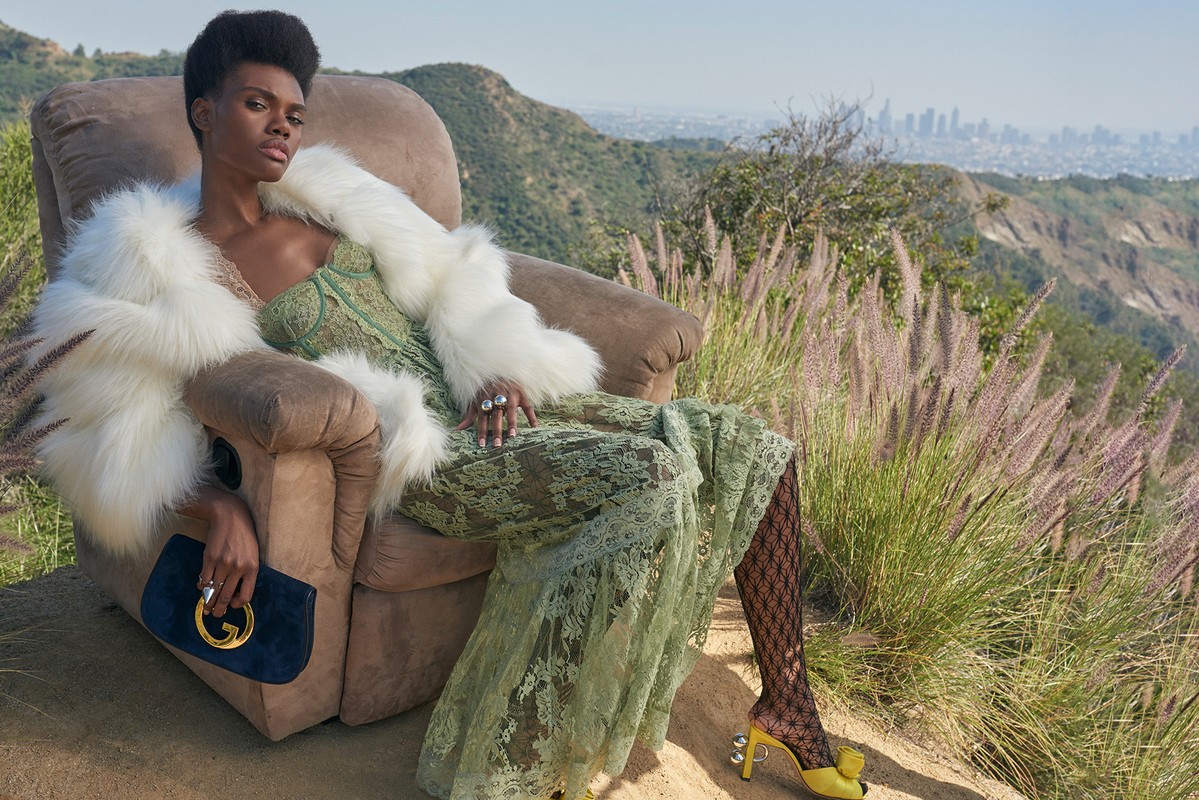 Elegant woman in a green dress and fur coat sitting in an armchair outdoors with city view, holding a dark blue Gucci purse.