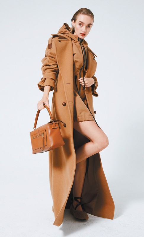 Elegant woman in a caramel-colored coat and dress holding a brown leather handbag against a clean white background in a studio setting.