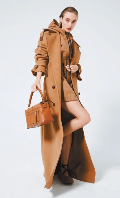 Elegant woman in a caramel-colored coat and dress holding a brown leather handbag against a clean white background in a studio setting.