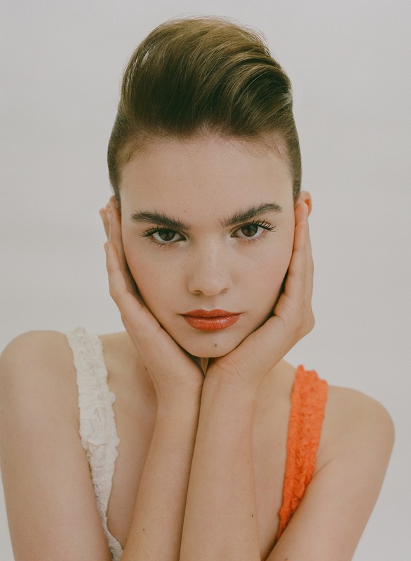 Close-up portrait of a young woman framing her face with hands, wearing white and orange tops against a plain background