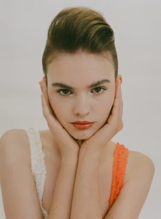 Close-up portrait of a young woman framing her face with hands, wearing white and orange tops against a plain background