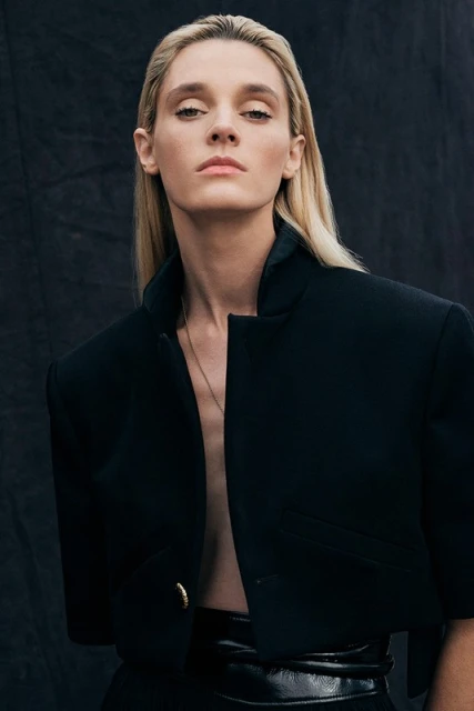 Portrait of a fair-skinned, blonde adult woman in a black crop jacket with a neutral facial expression against a dark backdrop.