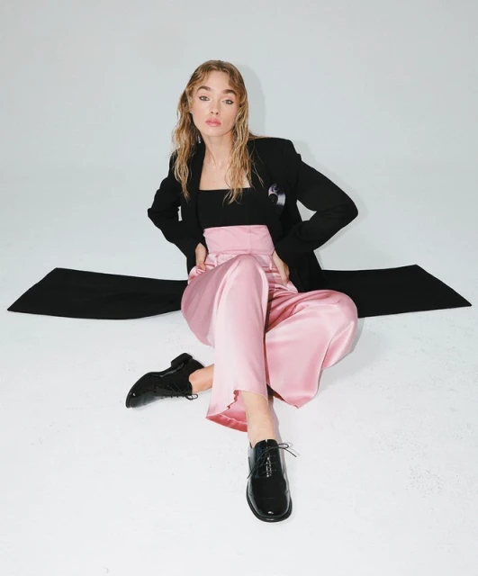 Young woman poses in black blazer, black top, pink pants and black patent-leather shoes against a white backdrop in a studio shot.